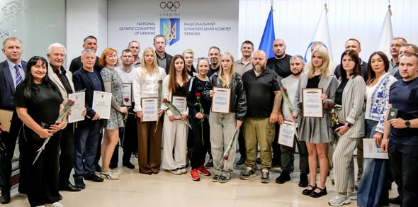 На зображенні велика група чоловіків та жінок позує для спільного фото в офіційному приміщенні на тлі стіни з логотипом та написами «Національний олімпійський комітет України», тримаючи в руках дипломи, грамоти та поодинокі квіти троянд.