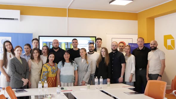 Група людей різного віку стоїть разом у світлій офісній кімнаті з жовтими елементами стін перед великим екраном та столами, позуючи для спільного офіційного фото
