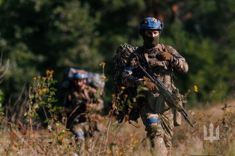 Двоє українських військовослужбовців у камуфляжному екіпіруванні, шоломах із синьою стрічкою та зі зброєю в руках рухаються гуськом крізь зарості сухої трави та квітів на тлі розмитого зеленого лісу.