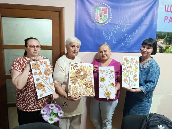 На зображенні четверо усміхнених жінок різного віку стоять поруч і тримають у руках свої хендмейд-картини з рослинними мотивами, створені з винних заторів та природних матеріалів, на тлі синього прапора з написом «Луганщина це Європа».
