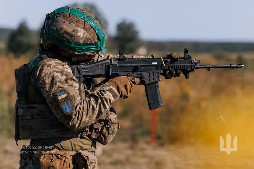 На зображенні показано українського військового у камуфляжній формі та шоломі з кавер-стрічкою, який цілиться і стріляє з автоматичної гвинтівки CZ BREN 2, під час чого з гвинтівки вилітає стріляна гільза.