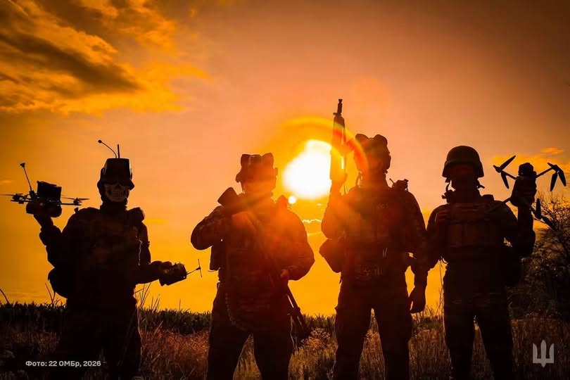 На зображенні чотири силуети українських військових із безпілотниками та зброєю стоять у полі на тлі яскравого помаранчевого заходу сонця.