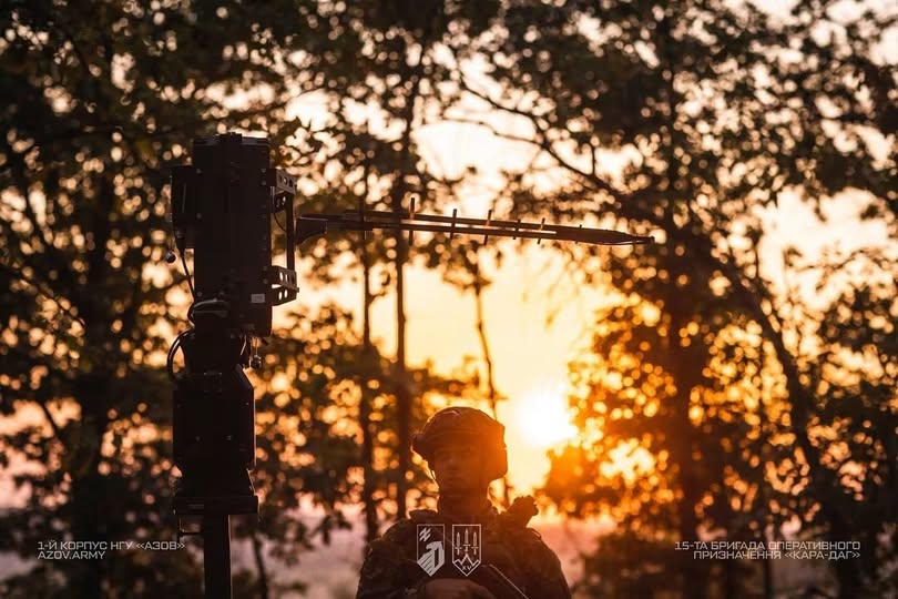 На тлі яскравого заходу сонця та силуетів дерев зображено військовослужбовця в касці, поруч із яким на передньому плані встановлено спеціальну військову антену або обладнання зв'язку на стійці.
