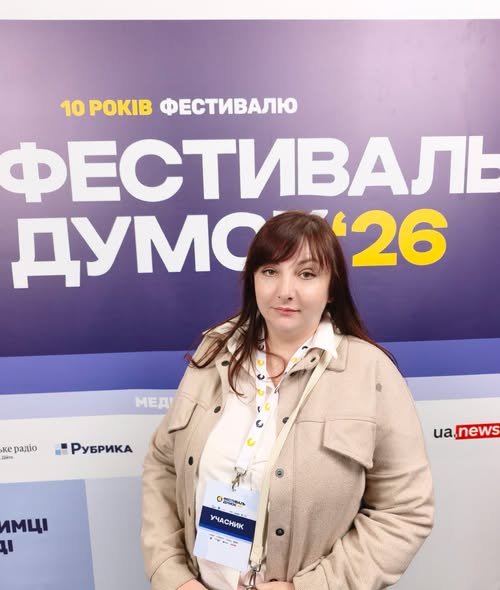 На зображенні темнокоса жінка середнього віку у світлій бежевій куртці та з бейджем «УЧАСНИК» на шиї стоїть у приміщенні на тлі фіолетового банера з великими написами «10 РОКІВ ФЕСТИВАЛЮ» та «ФЕСТИВАЛЬ ДУМОК '26».