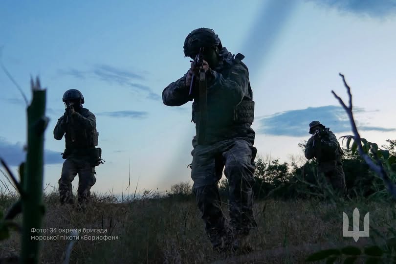 На зображенні на тлі вечірнього синього неба троє військовослужбовців у повному тактичному екіпіруванні та шоломах рухаються полем із сухою травою, тримаючи автомати нарівні очей та цілячись уперед, а в правому нижньому куті розміщено сірий малий герб України (тризуб).