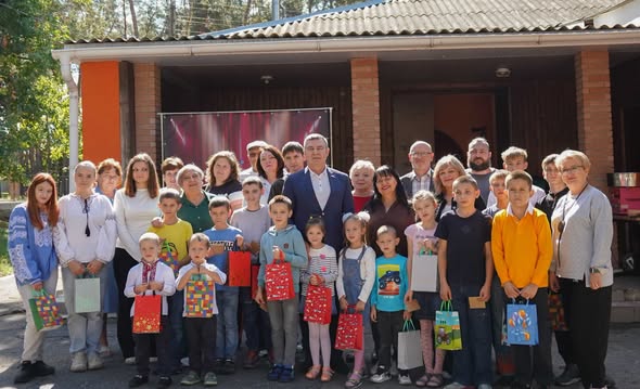 На тлі цегляного одноповерхового будинку та дерев просто неба стоїть велика група дітей різного віку та дорослих, деякі з яких тримають у руках подарункові пакети.