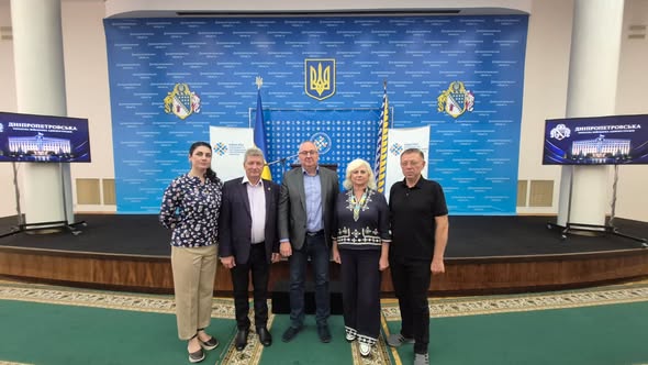 На сцені з офіційним синім тлом та гербом України стоять у ряд п'ятеро людей — три чоловіки та дві жінки.