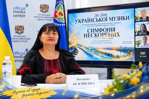 Катерина Безгинська у вишиванці сидить за столом на тлі прапора України та банера Луганської ОВА, а праворуч від неї розташовано великий екран із презентацією проєкту «Симфонія нескорених» до До Дня української музики та віконцями учасників онлайн-трансляції.