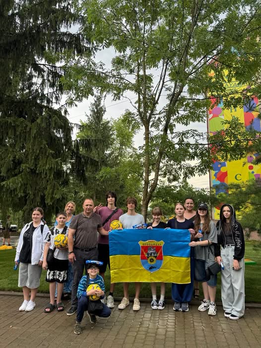 Група дітей та дорослих позує для фотографії на брукованій доріжці в парку, тримаючи в руках футбольні м'ячі та розгорнутий синьо-жовтий прапор із гербом міста Покровськ на тлі високих дерев і яскраво розфарбованого будинку.