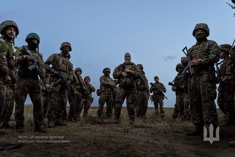 На панорамному фото на тлі сутінкового неба зафіксовано групу українських військовослужбовців у повному бойовому спорядженні зі зброєю в руках, які стоять півколом у полі, а в кутках розміщено напис «Фото: 34 окрема бригада морської піхоти «Борисфен»» та білий малий герб України.