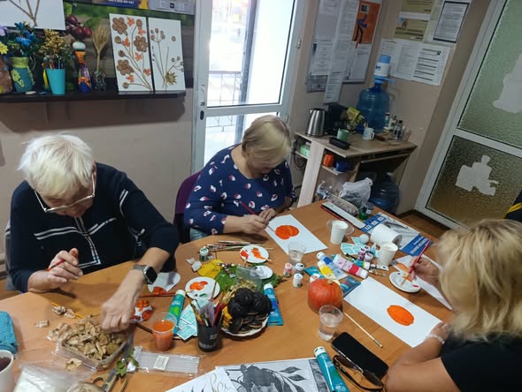 На зображенні три жінки старшого віку сидять за дерев'яним столом у світлій кімнаті та захоплено займаються творчістю, малюючи фарбами на папері та викладаючи композиції з природних матеріалів.