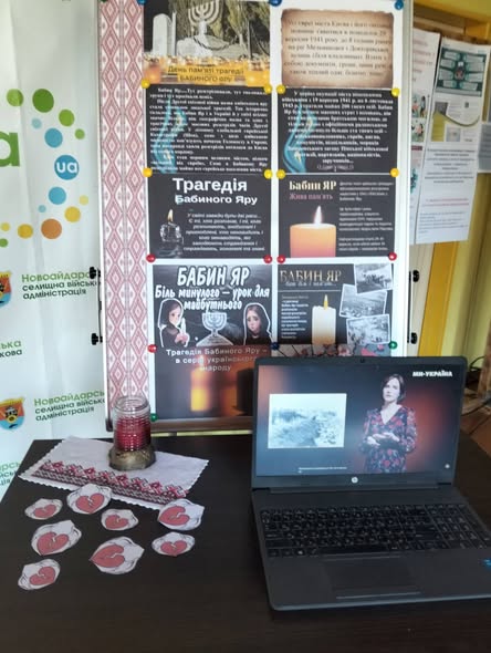 На зображенні на столі стоїть відкритий ноутбук, на екрані якого транслюється відео про Бабин Яр, а поруч лежать паперові серця, вишита серветка зі свічкою та високий інформаційний стенд Новоайдарської селищної військової адміністрації з плакатами про трагедію Бабиного Яру.