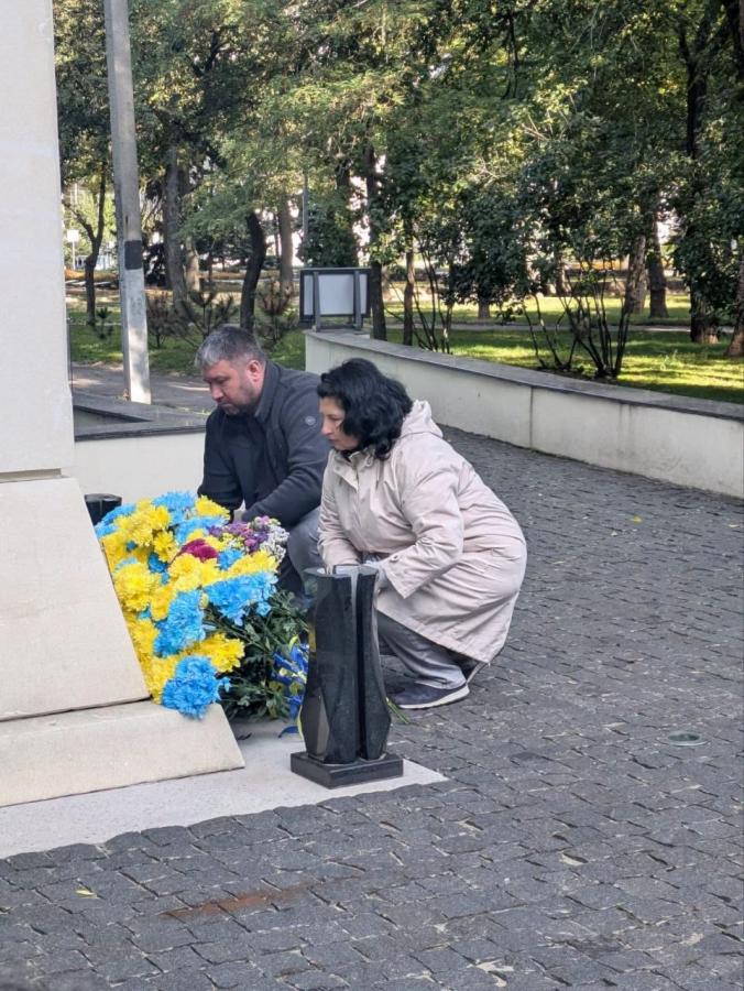 На світлині чоловік у темній куртці та жінка у бежевому пальті присіли біля підніжжя світлого кам'яного монумента, щоб покласти великий букет із жовтих і блакитних квітів на знак вшанування пам'яті.