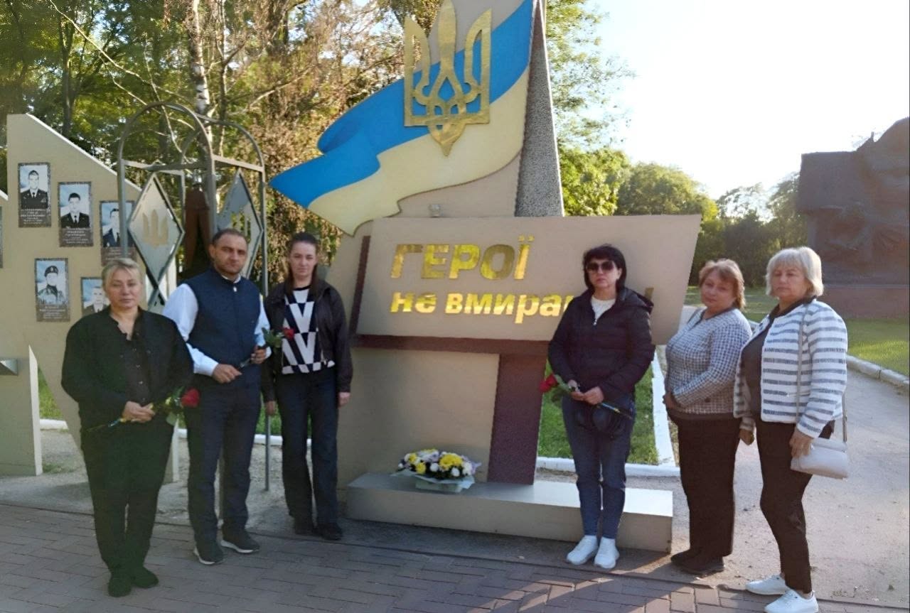 На світлині група з шістьох людей стоїть на вулиці біля меморіального пам'ятника з написом «Герої не вмирають», який увінчаний гербом та прапором України.