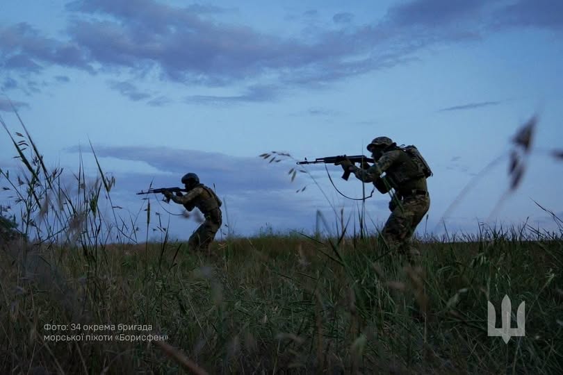На зображенні двоє українських військових у камуфляжі та шоломах зі зброєю в руках пересуваються полем серед високої трави у сутінках під хмарним небом.