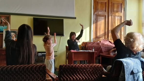 У просторій залі з великими вікнами та розписними стінами жінка за невеликим столом веде зустріч із групою людей різного віку, які сидять навколо на кріслах і диванах та разом повторюють жест, склавши долоні перед собою.