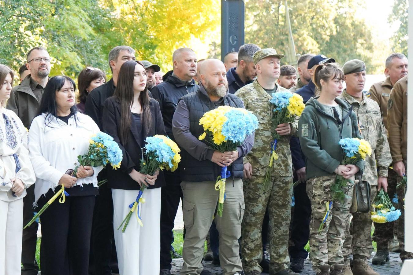На світлині велика група людей, серед яких цивільні та військові у камуфляжі, стоять разом на вулиці під час урочистого заходу, тримаючи в руках букети жовтих і блакитних квітів.