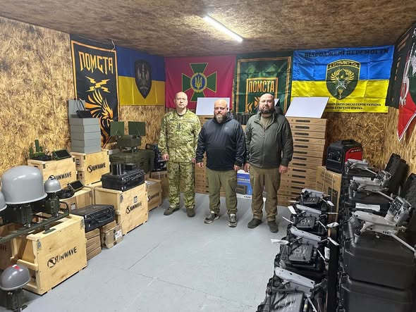 Три чоловіки — один у військовій формі та двоє у цивільному одязі — стоять у приміщенні військового складу, оточені коробками з обладнанням, дронами та прапорами підрозділу «Помста» на стінах.