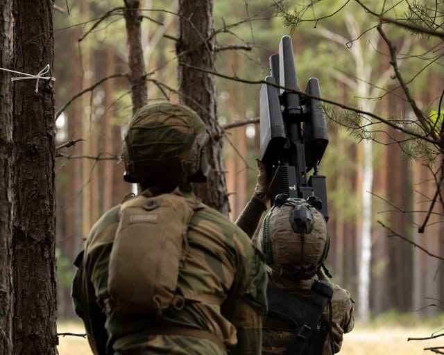 На зображенні двоє військових у камуфляжній формі та шоломах стоять у густому сосновому лісі, при цьому один із них тримає в руках спрямовану вгору велику чорну антидронову рушницю.