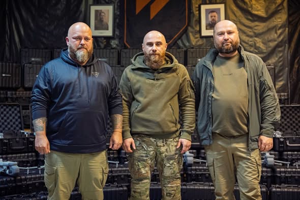 Три бородаті чоловіки у військовому та тактичному одні đứng пліч-о-пліч на тлі військового спорядження, ящиків та портретів на стіні.