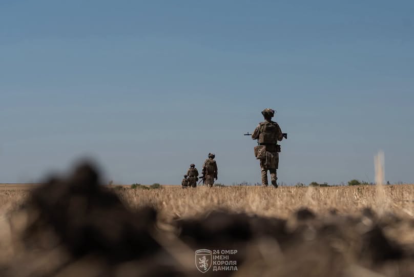 На фотографії ракурсом знизу зображено чотирьох українських військовослужбовців у повному екіпіруванні зі зброєю, які віддаляються ланцюжком по сухому полю на тлі чистого блакитного неба, а знизу розміщено логотип «24 ОМБр імені короля Данила».