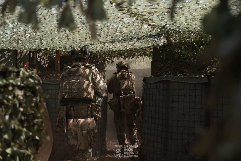 Двоє військовослужбовців у повному бойовому спорядженні стоять спиною до камери всередині облаштованого окопу під маскувальною сіткою.