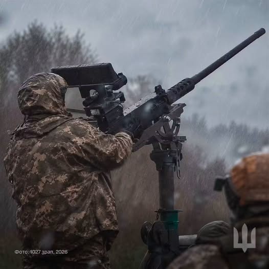 На фотографії в похмуру дощову погоду зображено військовослужбовця в камуфляжній куртці з капюшоном зі спини, який керує встановленим на турелі великокаліберним кулеметом із тепловізійним або оптичним прицілом, спрямованим угору на тлі дерев.