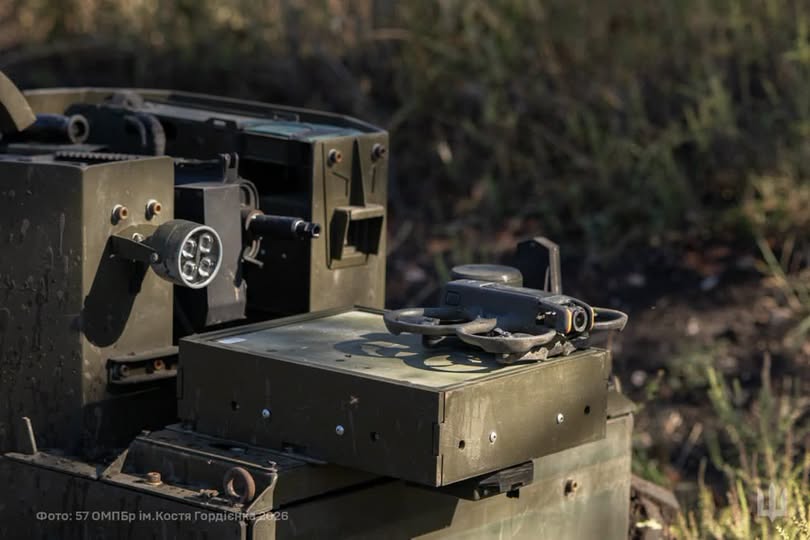 Фотографія військової техніки захисного кольору на тлі сухої трави, на металевому корпусі якої лежить невеликий безпілотник типу квадрокоптер.