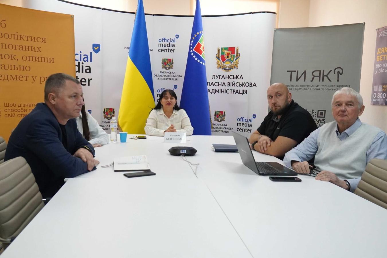 Група людей сидить за великим білим Т-подібним столом під час офіційного заходу в Луганській обласній військовій адміністрації, на тлі державного прапора України та брендованих банерів «official media center» та «Ти як?»
