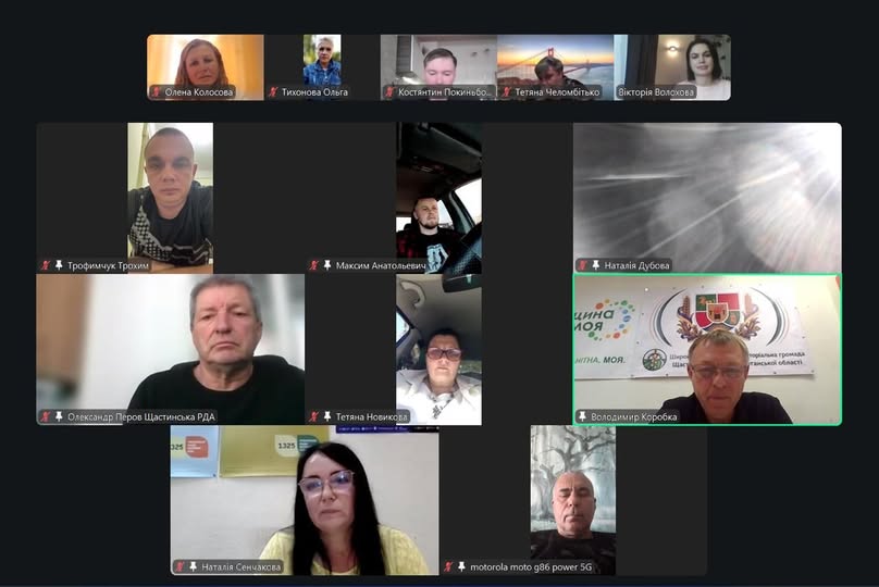 Скриншот онлайн-наради на платформі Zoom або Google Meet демонструє вікно відеоконференції з одинадцятьма вікнами учасників, серед яких видно чоловіків та жінок у домашніх умовах або в автомобілях, підписаних українськими іменами (зокрема Олександр Перов Щастинська РДА, Володимир Коробка на тлі офіційного банера громади, Наталія Сенчакова та інші), які слухають або беруть участь у спілкуванні.
