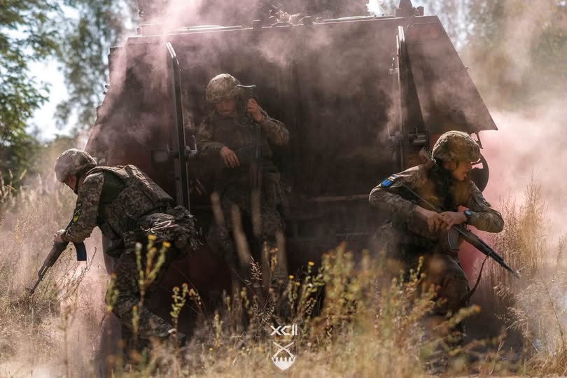 Трое украинских военных в полной боевой экипировке и шлемах стремительно десантируются из открытых задних дверей бронетранспортера через густую завесу розового дыма на заросшее травой поле.