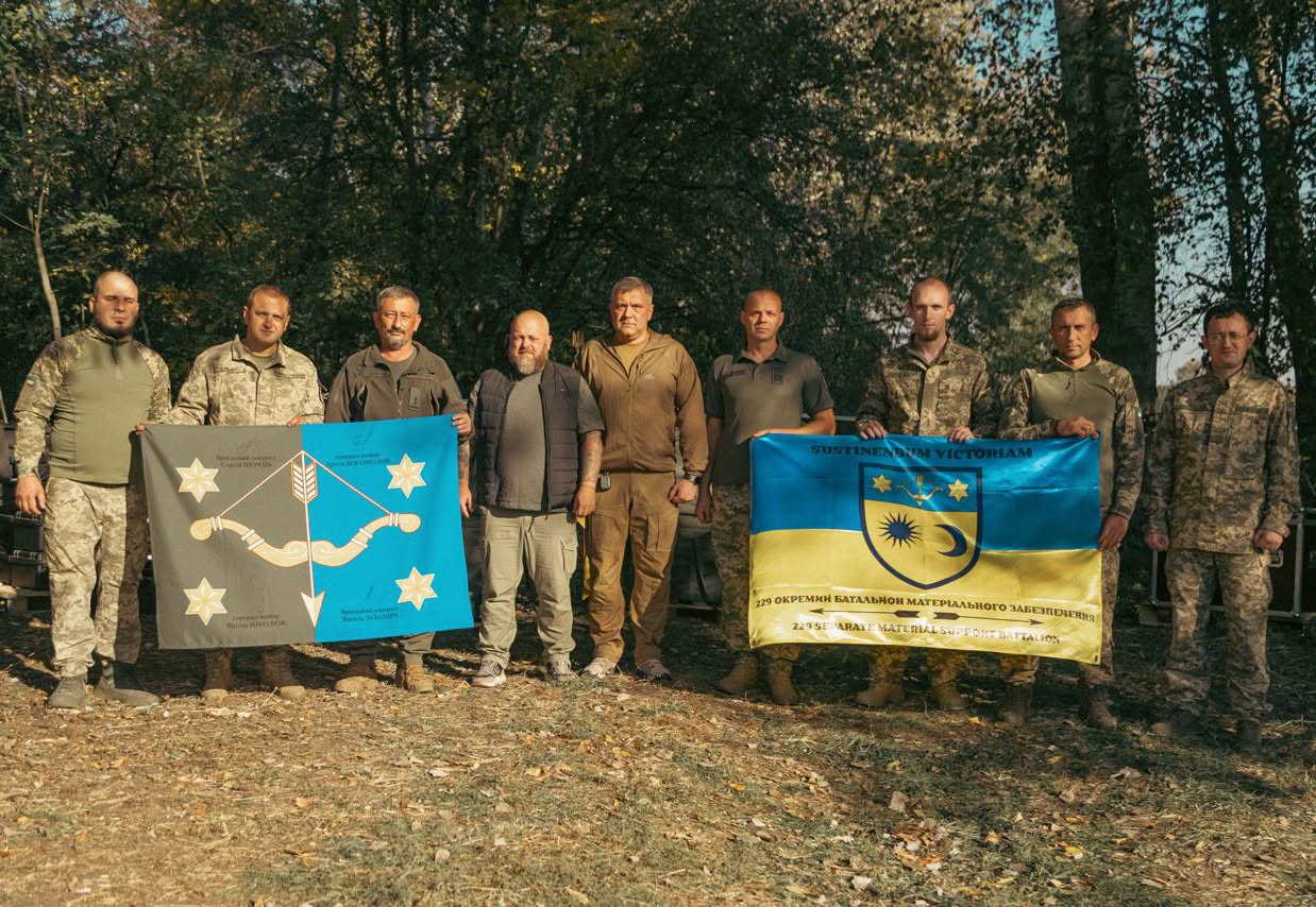 На горизонтальному фото на тлі осінніх дерев і сухого листя стоять дев'ятеро чоловіків, здебільшого у військовій формі Збройних Сил України, які тримають два прапори: ліворуч — сіро-блакитне знамено з білим натягнутим луком і чотирма зірками, а праворуч — синьо-жовтий прапор із гербом та написом «229 окремий батальйон матеріального забезпечення» українською та англійською мовами