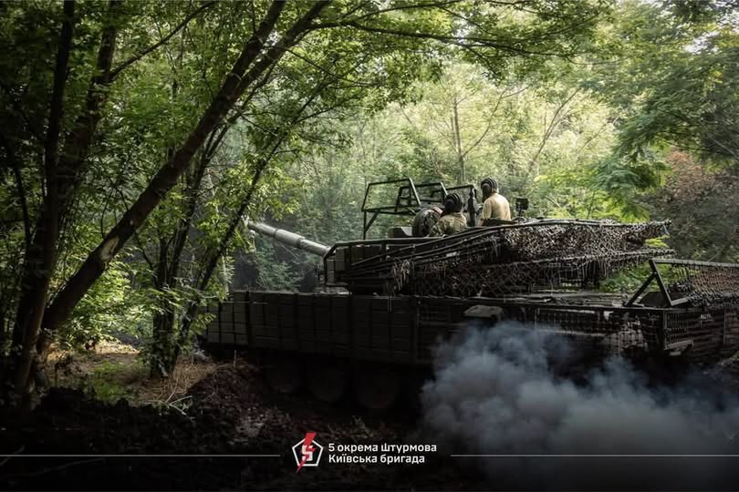 На фото зображено танк Сухопутних військ ЗСУ з екіпажем, який маневрує серед густих зелених дерев у лісистій місцевості, залишаючи за собою клуби густого сірого диму, а знизу розміщено логотип та напис «5 окрема штурмова Київська бригада».