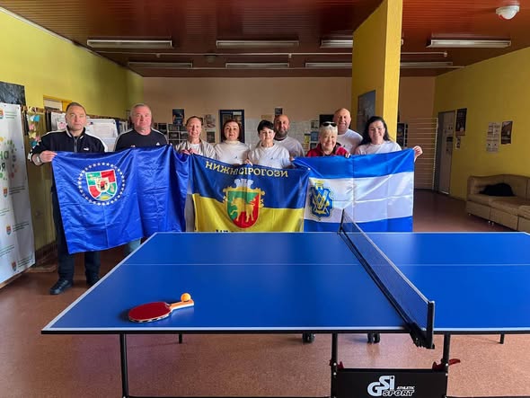Група чоловіків та жінок стоїть у приміщенні за синім тенісним столом, на якому лежить ракетка з м'ячиком, та тримає три прапори, серед яких жовто-блакитний прапор із гербом та написом «Нижні Сірогози».