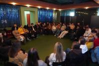На знімку зафіксовано групову зустріч великої кількості людей (близько 20-25 осіб), які сидять у великому колі або півколі. Ракурс обраний зверху та ззаду від частини присутніх, що створює ефект залученості глядача в середину події. Центр кімнати залишається вільним, що підкреслює формат відкритого діалогу або спільного обговорення.