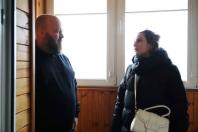 На фотографії зображено чоловіка з бородою в темному одязі та жінку в чорній куртці з білою сумкою, які стоять обличчям один до одного в приміщенні з дерев'яною обшивкою та великими вікнами, ймовірно, під час розмови