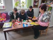 Чотири дитини та доросла жінка, ймовірно, вчителька чи бібліотекарка, сидять навколо столу та займаються творчістю, використовуючи папір, клей та олівці в приміщенні, схожому на бібліотеку чи клас. Цей альтернативний текст описує основну сцену, включаючи кількість людей, місце дії та предмети, які вони використовують.