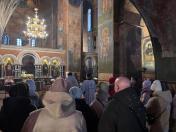 Група людей у зимовому одязі, включаючи куртки з хутряними комірами та шапки, зібралася в приміщенні, схожому на церкву або храм, про що свідчать розташовані на стінах релігійні ікони та інформаційні стенди.