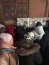 Група людей у зимовому одязі, включаючи куртки з хутряними комірами та шапки, зібралася в приміщенні, схожому на церкву або храм, про що свідчать розташовані на стінах релігійні ікони та інформаційні стенди.