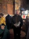 Чоловік у чорній куртці та окулярах стоїть у приміщенні церкви з історичними фресками на стінах, тримаючи в руках пакет із ймовірними свічками, тоді як інші парафіяни та священник у жовтому вбранні знаходяться на задньому плані.