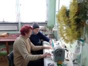 Дві жінки з рудим волоссям у світлому светрі та в сірій шапці в синьому одязі сидять за столом зі швейною машиною, оточені вікном, кімнатними рослинами та новорічними прикрасами.
