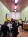 Кілька людей працюють за столами з комп’ютерами та документами в офісному приміщенні, де видно українські прапори.