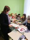 Кілька людей, включаючи двох жінок на передньому плані, зібралися навколо столу в кімнаті для рукоділля з різними матеріалами, малюнками, м'якими іграшками та іншими людьми на фоні