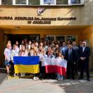 Група людей, включно з учнями та дорослими представниками адміністрації Bursa Szkolna im. Janusza Korczaka W Chełmie, позує біля будівлі з українським та польським прапорами.