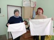 Дві жінки в повсякденному одязі стоять у приміщенні з шафами та іншими предметами рукоділля на фоні та тримають білі тканинні вироби, схожі на мішки або наволочки.