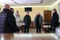 Група людей у верхньому одязі (чоловіки в темних куртках і жінка в світлому пальті) стоїть у приміщенні з табличкою «ВИХІД» над дерев'яними дверима.