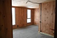 Пуста кімната з незавершеним ремонтом має стіни та стелю, обшиті дерев’яними панелями, бетонну підлогу, два високих вузьких вікна та видимі електричні кабелі