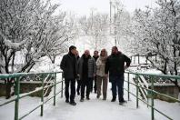 Кілька людей, включаючи чоловіків у темних куртках і жінку в світлому пальті та шарфі, йдуть засніженим мостом із зеленими металевими перилами, оточеним деревами, вкритими снігом.