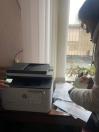 Людина в окулярах і світлій куртці працює з багатофункціональним принтером HP, тримаючи в руках круглий предмет, а на столі поруч розкидані папери.