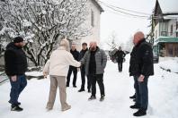 Кілька чоловіків у зимовому одязі стоять на засніженій вулиці перед будинками та деревами, при цьому двоє центральних чоловіків тиснуть руки.